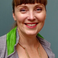 Podcast guest, Ichsani Wheeler, wearing a gray plaid jacket with a bright green collar and a gold necklace, standing against a plain blue-gray background.