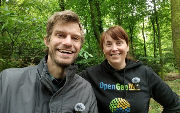 Two people stand side by side in a lush, green forest. The person on the left is wearing a black "OpenGeoHub" hoodie with a colorful globe logo and a microphone clipped to the chest. The person on the right is wearing a grey jacket with a microphone attached. Both are facing the camera, surrounded by tall trees and dense foliage.