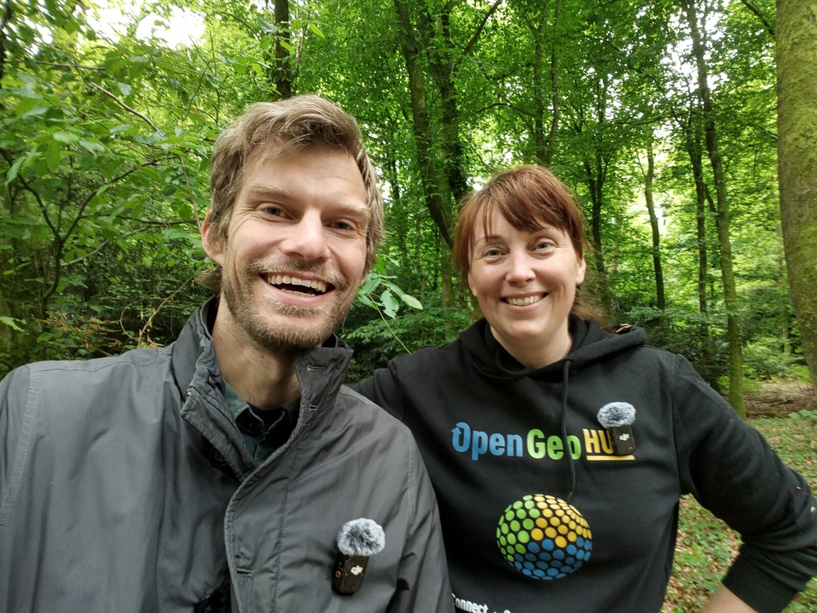Two people stand side by side in a lush, green forest. The person on the left is wearing a black "OpenGeoHub" hoodie with a colorful globe logo and a microphone clipped to the chest. The person on the right is wearing a grey jacket with a microphone attached. Both are facing the camera, surrounded by tall trees and dense foliage.