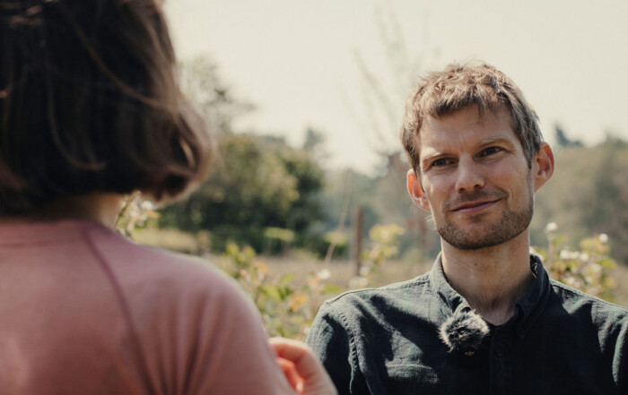 Two people are outdoors having a conversation. One person, wearing a dark shirt with a microphone clipped to the collar, is facing the camera. The other person, with shoulder-length brown hair and wearing a pinkish shirt, is seen from behind. The background features greenery and a soft, natural light, indicating a relaxed, outdoor setting