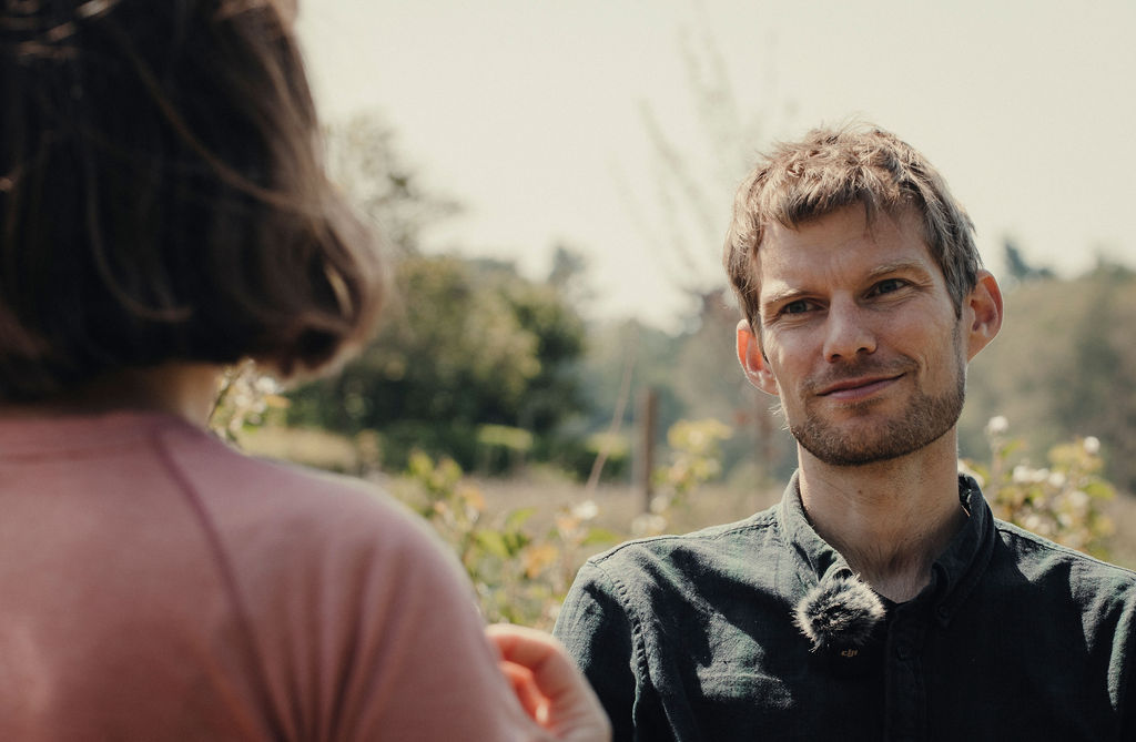Two people are outdoors having a conversation. One person, wearing a dark shirt with a microphone clipped to the collar, is facing the camera. The other person, with shoulder-length brown hair and wearing a pinkish shirt, is seen from behind. The background features greenery and a soft, natural light, indicating a relaxed, outdoor setting