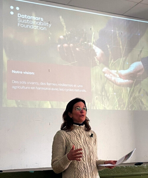 A woman in a cream cable-knit sweater speaks in front of a projected Datamars Sustainability Foundation slide showing hands holding soil and French text about living soils and resilient farms.
