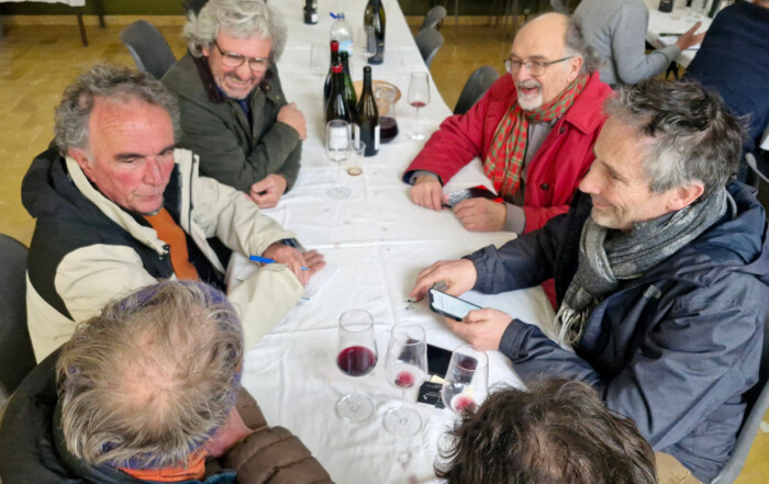 Winegrowers sit closely around a long table during a breakout discussion, with notebooks, smartphones, and half-filled glasses of red wine in front of them, inside a simple community hall.