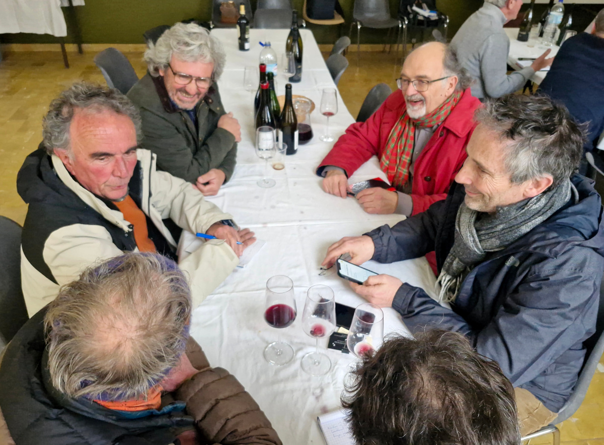 Winegrowers sit closely around a long table during a breakout discussion, with notebooks, smartphones, and half-filled glasses of red wine in front of them, inside a simple community hall.